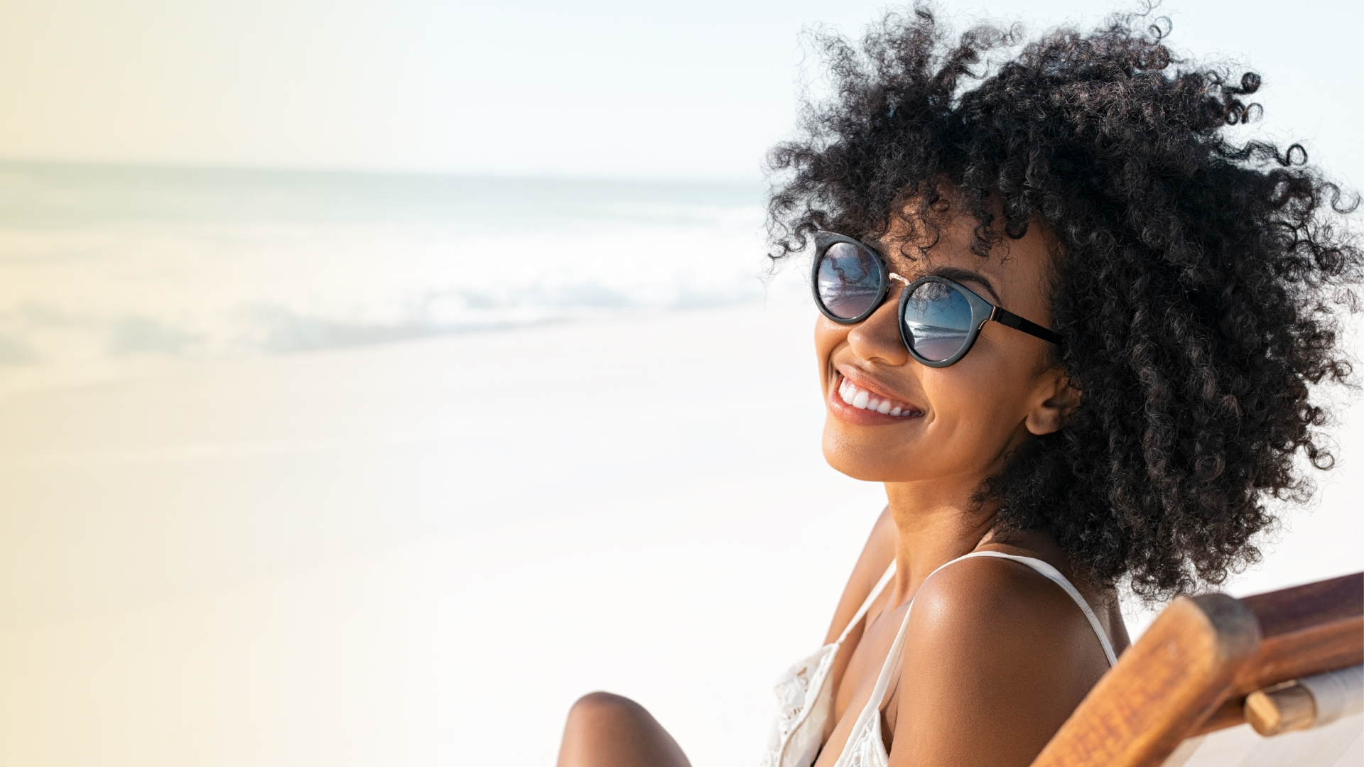 Smiling woman with curly hair and sunglasses sits on a beach chair, enjoying a sunny day by the ocean. The scene conveys happiness and relaxation.