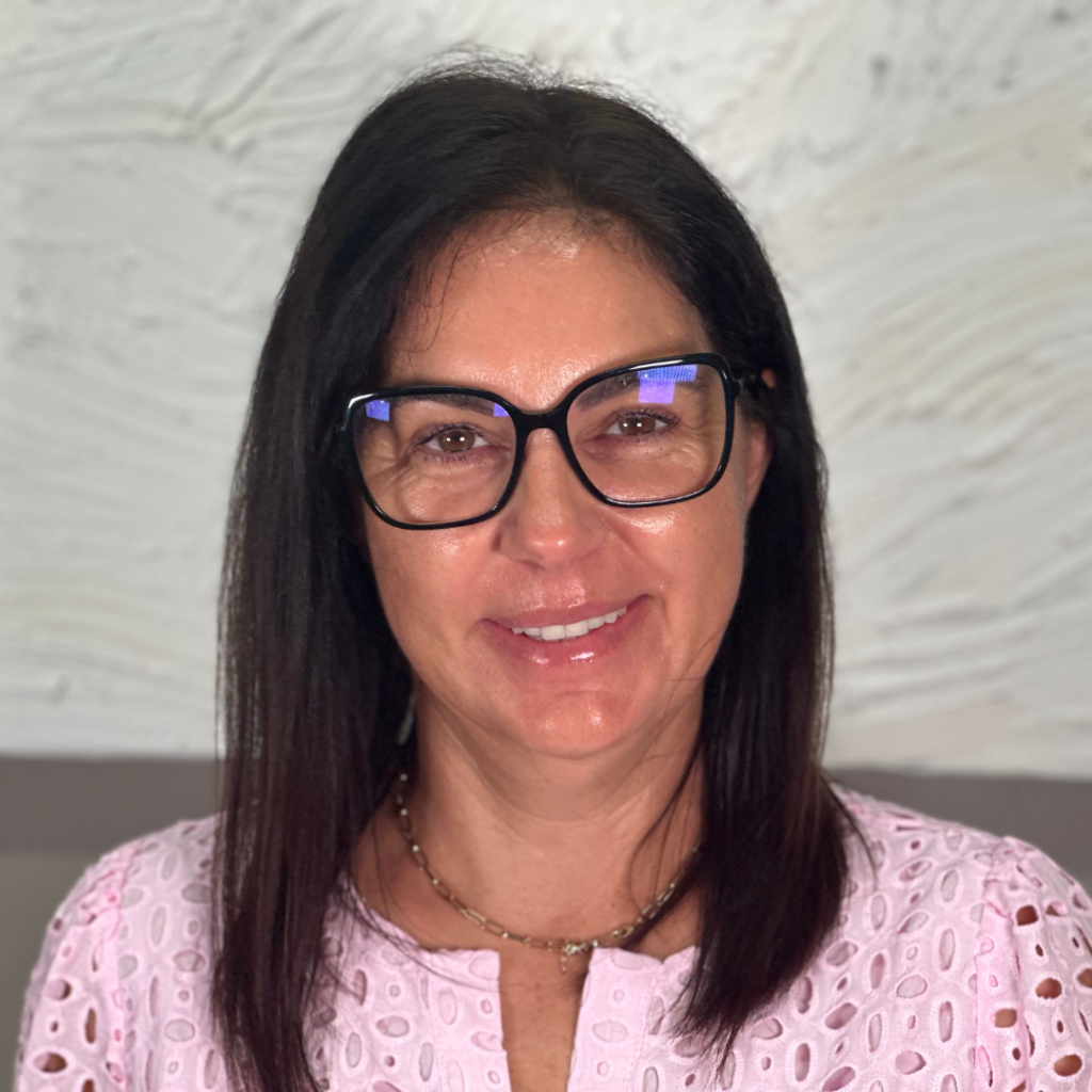 A woman with straight dark hair, wearing black-rimmed glasses and a light pink eyelet blouse, is smiling at the camera. The background is a textured, off-white wall.