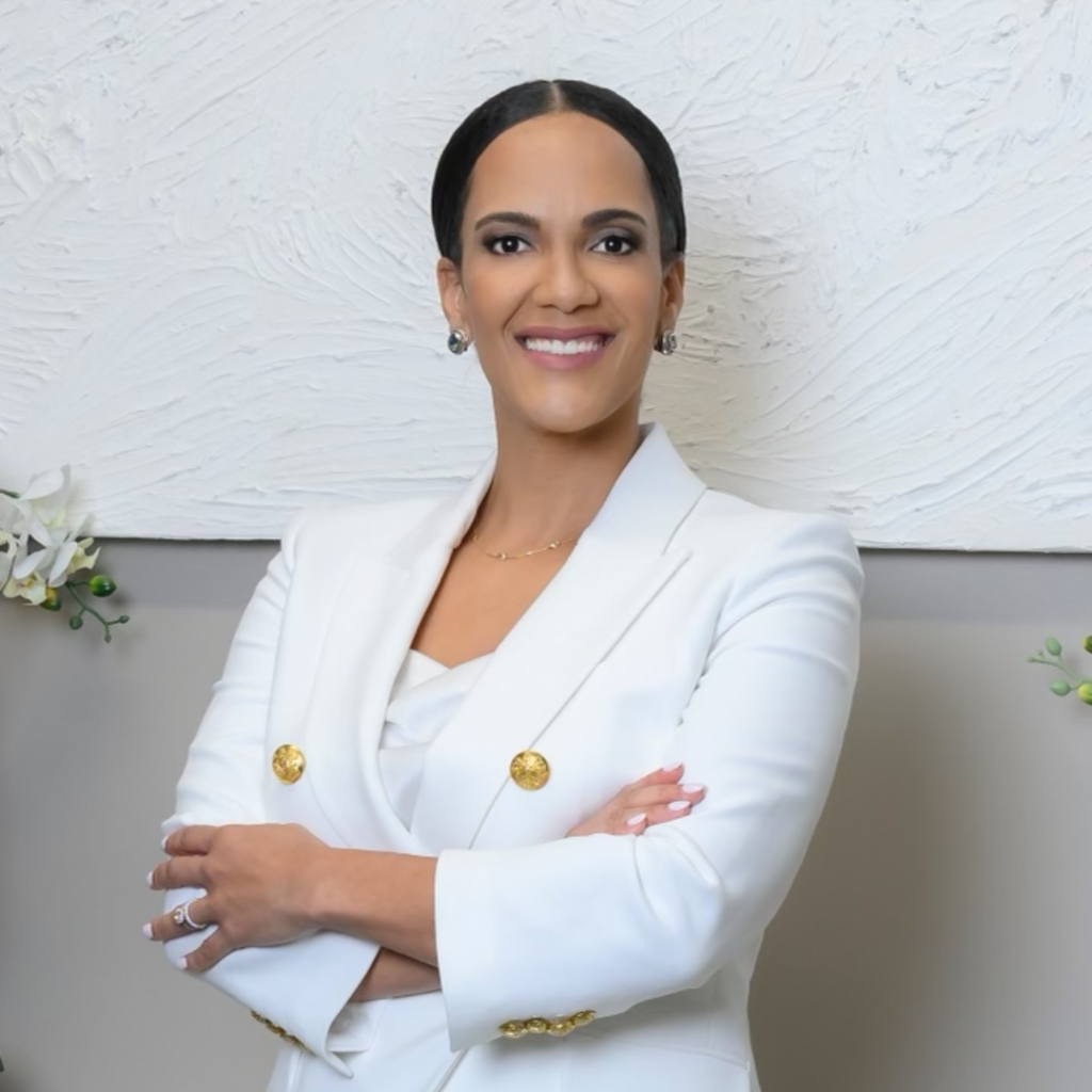 A dentist in a white blazer and light blue pants stands indoors, smiling, with one hand resting on a countertop beside a potted white orchid. The background features grey walls and abstract art.