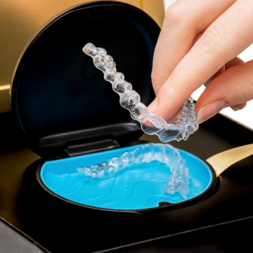 A hand places a clear Invisalign dental aligner into a blue tray inside a black and gold case, with another aligner already in the tray.