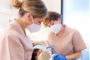 Two female dental professionals at a Dental Clinic, wearing masks and gloves, perform a dental procedure on a patient lying in the chair. One uses dental tools while the other assists.