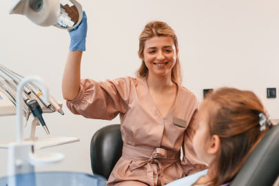 A smiling dentist in a peach coat at the dental clinic adjusts an overhead light while talking to a young patient sitting in a dental chair. Dental tools are visible nearby.
