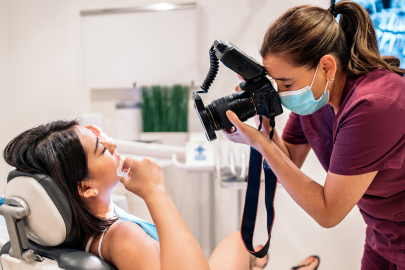 A dental professional at a dental clinic, wearing scrubs and a mask, takes close-up photos of a patient’s mouth as she sits in the dental chair with her mouth open.