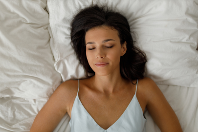 A woman with dark hair lies on her back in bed with white sheets, eyes closed and appearing peaceful—perhaps dreaming of her recent visit to the dental clinic. She is wearing a light-colored camisole top.
