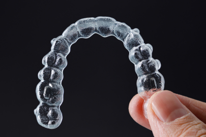 A hand holding a clear dental aligner against a dark background. The transparent aligner, shaped to fit over teeth, is commonly provided by a dental clinic for orthodontic treatments.