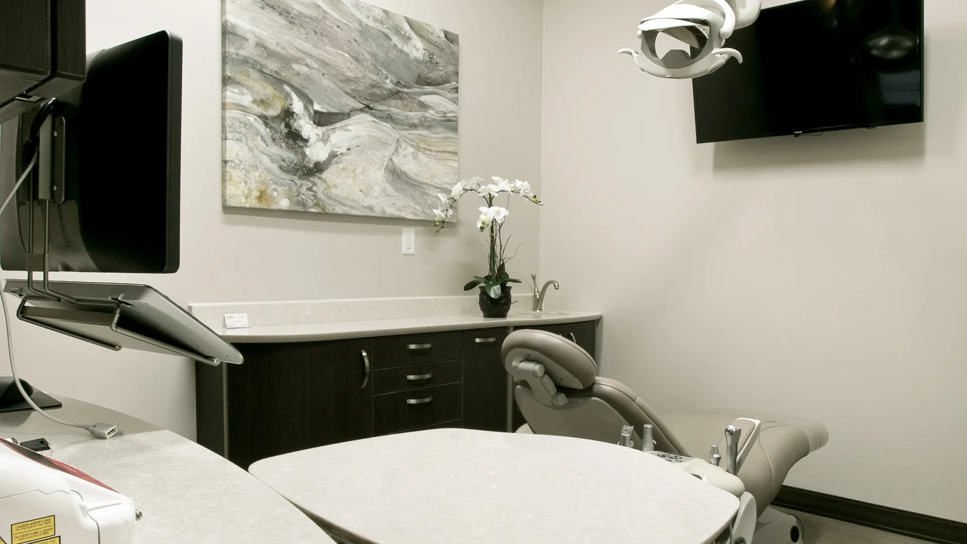A modern dental care office featuring a reclining examination chair, a mounted TV, computer monitors, a marble countertop, dark wood cabinets, an abstract wall painting, and a white orchid in a vase.