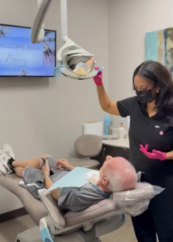 A dental professional in black scrubs and pink gloves adjusts an overhead light for a patient reclining in a dental chair. The atmosphere is calm and welcoming.