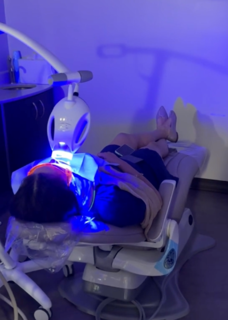 A dentist, wearing gloves and a mask, examines a patient's teeth using dental instruments. The patient lies on a chair under bright lights, conveying a clinical atmosphere.