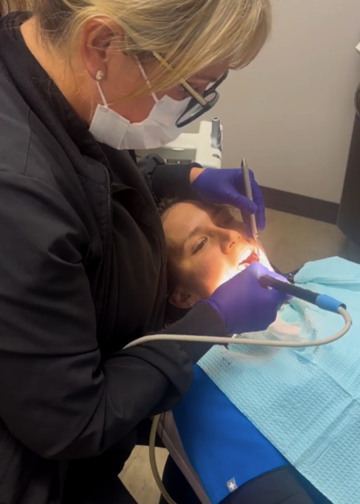 A dentist, wearing gloves and a mask, examines a patient's teeth using dental instruments. The patient lies on a chair under bright lights, conveying a clinical atmosphere.