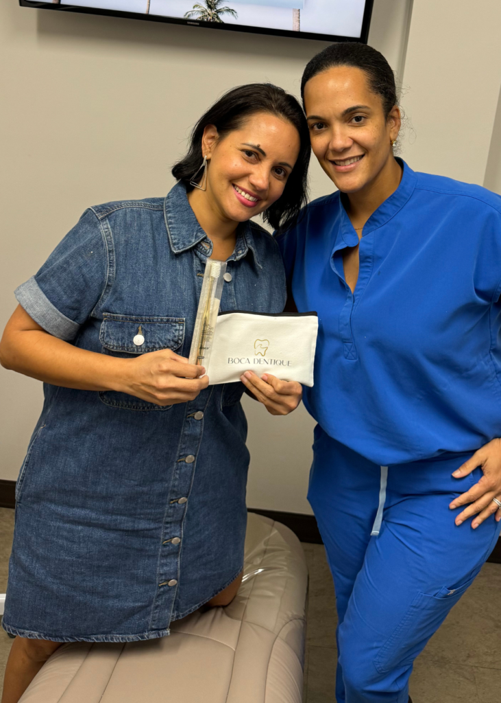 Two women smiling in a dental office setting. One wears a denim dress, holding dental care items; the other in blue scrubs stands beside her.