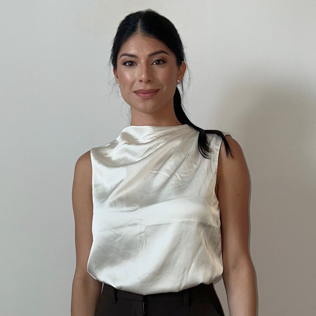 A woman with long dark hair in a low ponytail is standing and smiling. She is wearing a sleeveless, silky white top and dark pants, representing the Boca Dentique Team against a plain light-colored background.