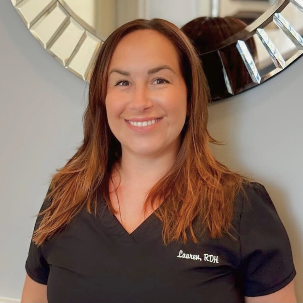 A woman with long brown hair, wearing black scrubs embroidered with Lauren, RDH, stands smiling in front of a decorative circular mirror on a light-colored wall as part of the Boca Dentique Team.