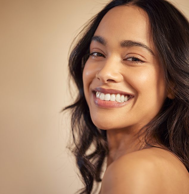 A woman with long, wavy dark hair smiles brightly at the camera against a soft beige background. She has glowing skin and appears happy and confident.