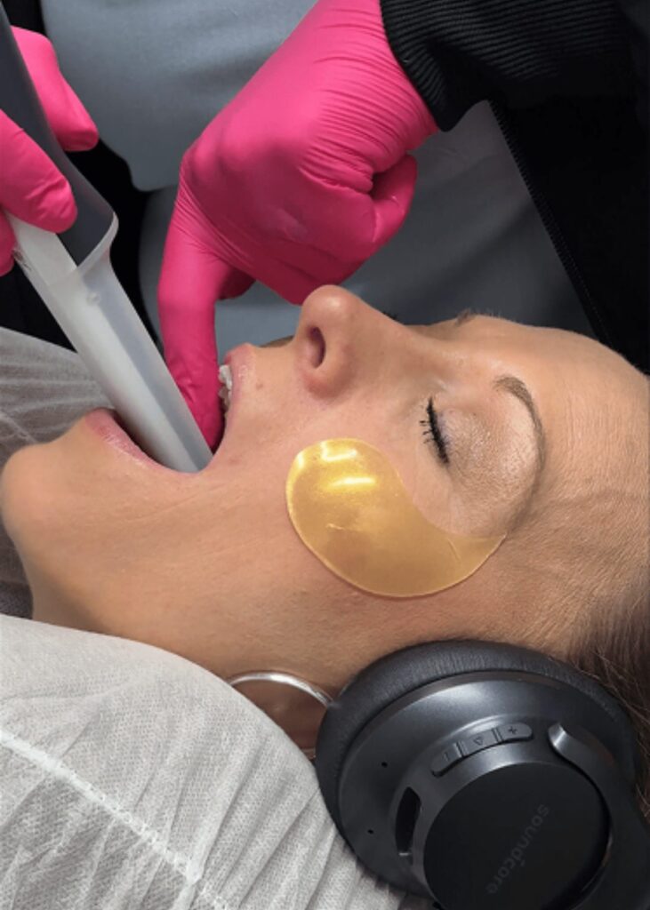 A woman wearing headphones and a gold under-eye patch receives a Dental Cleaning. A gloved hand holds a dental tool in her mouth as she rests on the treatment chair, eyes closed and relaxed.