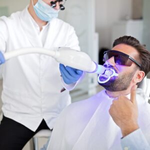 A man in sunglasses gives a thumbs up while receiving teeth whitening from a dentist using a blue light device. The tone is positive and professional.